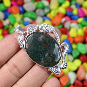 Puede incluir: Un colgante de forma ovalada con una piedra preciosa de ágata musgosa pulida con inclusiones de color verde oscuro, engastada en un marco de metal de tono plateado. El marco muestra intrincados adornos y patrones de hojas.