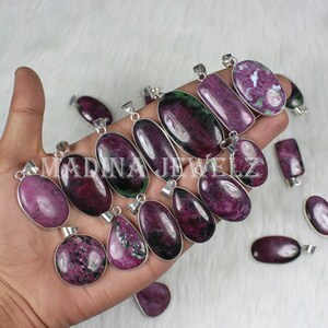 以下が含まれることがあります： シルバーカラーのセッティングが施されたルビーペンダントのコレクション。ペンダントは、楕円形、ティアドロップ型、長方形など、さまざまな形状をしており、濃い赤色と緑色の色合いが混ざっています。ペンダントは白い表面に表示されています。