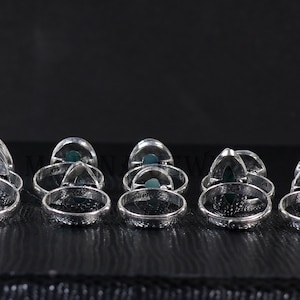 Pode incluir: Uma cole&ccedil;&atilde;o de an&eacute;is de prata com uma pedra preciosa verde triangular embutida em uma banda decorativa. Os an&eacute;is est&atilde;o dispostos em uma fileira sobre uma superf&iacute;cie escura, mostrando seu design intrincado e acabamento polido. Cada anel &eacute; marcado com "925".