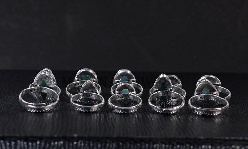 Pode incluir: Uma cole&ccedil;&atilde;o de an&eacute;is de prata com uma pedra preciosa verde triangular embutida em uma banda decorativa. Os an&eacute;is est&atilde;o dispostos em uma fileira sobre uma superf&iacute;cie escura, mostrando seu design intrincado e acabamento polido. Cada anel &eacute; marcado com "925".