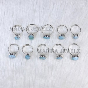 Pode incluir: Dez an&eacute;is de prata ajust&aacute;veis com engastes de pedras preciosas azul claro s&atilde;o exibidos em uma superf&iacute;cie branca e texturizada. Os an&eacute;is est&atilde;o dispostos em duas fileiras. O texto "MADINA JEWELZ" est&aacute; impresso na superf&iacute;cie.