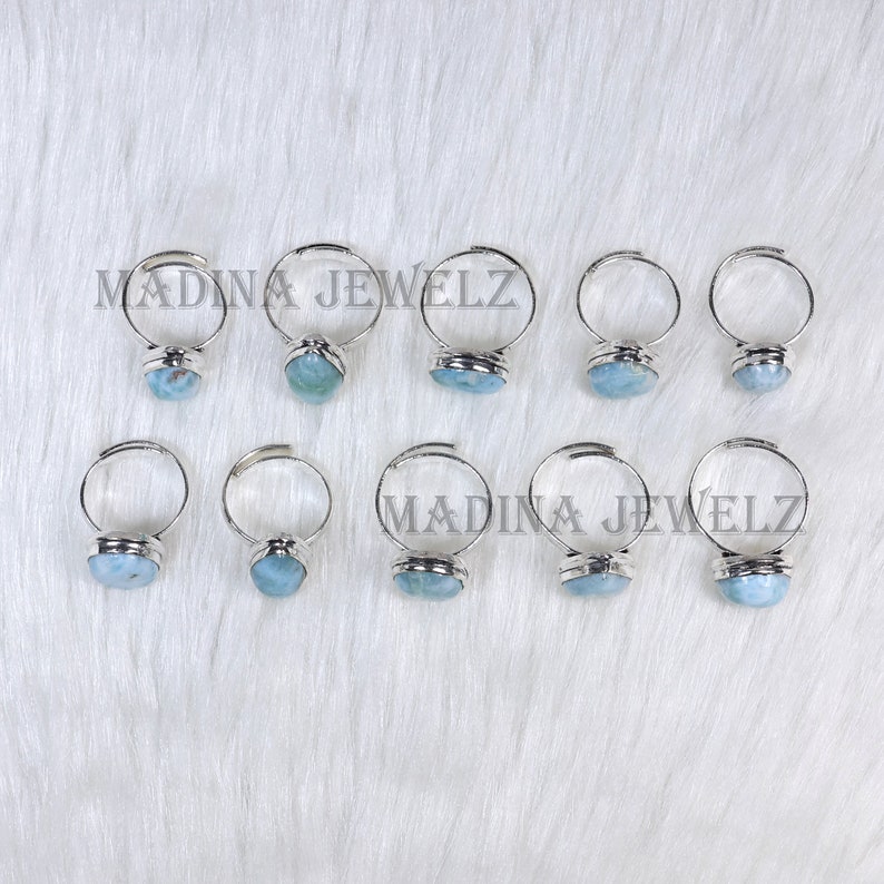 Pode incluir: Dez an&eacute;is de prata ajust&aacute;veis com engastes de pedras preciosas azul claro s&atilde;o exibidos em uma superf&iacute;cie branca e texturizada. Os an&eacute;is est&atilde;o dispostos em duas fileiras. O texto "MADINA JEWELZ" est&aacute; impresso na superf&iacute;cie.