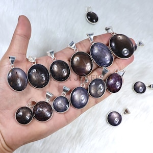 Może przedstawiać: Kolekcja owalnych wisiorków o ciemnym, opalizującym połysku. Każdy wisiorek osadzony jest w srebrnej oprawie. Kamienie szlachetne mają różne kolory, od głębokiego brązu po fiolet i szarość. Wisiorki są prezentowane na białej, teksturowanej powierzchni.