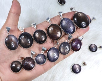 Puede incluir: Una colección de colgantes ovalados con un brillo oscuro e iridiscente. Cada colgante está engastado en un marco de metal plateado. Las piedras preciosas varían en color, desde marrón oscuro hasta púrpura y gris. Los colgantes se muestran sobre una superficie blanca texturizada.