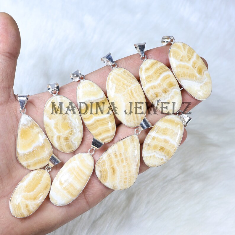 Pu&ograve; includere: Una collezione di pendenti con pietre preziose di varie forme ovali e a goccia. I pendenti presentano un motivo a righe crema e bianco, incastonati in metallo color argento. Il testo "MADINA JEWELZ" &egrave; visibile nell'immagine.