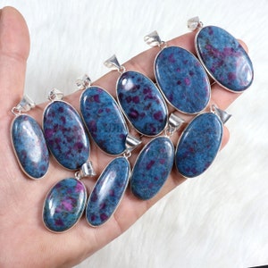 May include: A collection of nine oval-shaped blue and purple gemstone pendants with silver bail settings. The pendants are arranged in a hand, with the largest pendant at the top and the smallest pendant at the bottom.