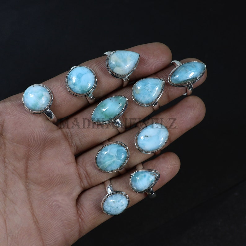 Pode incluir: Uma cole&ccedil;&atilde;o de an&eacute;is de prata esterlina com pedras preciosas de larimar azul claro em v&aacute;rias formas. Os an&eacute;is s&atilde;o exibidos em um fundo escuro, mostrando as pedras polidas e as configura&ccedil;&otilde;es de prata. As pedras preciosas t&ecirc;m uma apar&ecirc;ncia marmorizada.