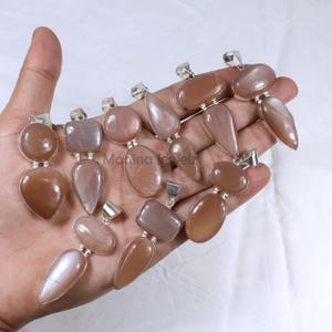 Puede incluir: Una colección de 12 colgantes de piedra de sol plateados, con una variedad de formas y tamaños. Los colgantes se muestran en una mano, mostrando su brillo y color únicos.