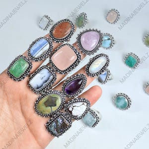 May include: A collection of gemstone rings with various shapes and colors, including green, pink, and purple stones. Each ring has a silver-colored band and a beaded border around the stone. The rings are displayed on a hand.