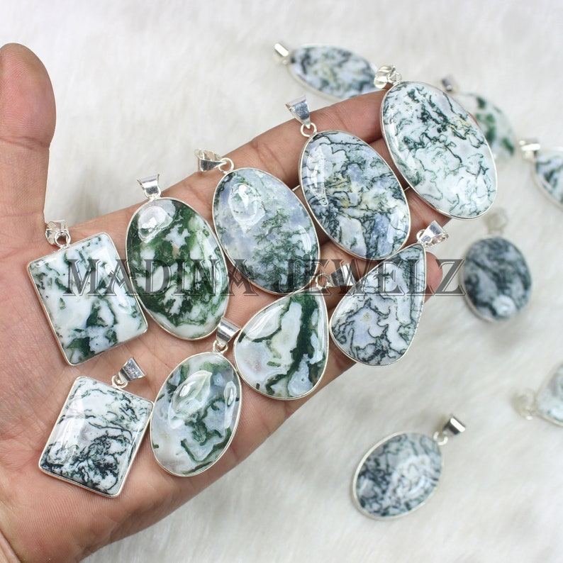 May include: A collection of silver-toned pendants with various shapes, featuring white and green moss agate stones. The pendants are displayed on a soft, white surface. The stones have unique, natural patterns.