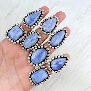 May include: A collection of silver-toned rings with various shapes, each featuring a light blue gemstone. The gemstones are set in a beaded border. The rings are displayed on a hand against a white background.