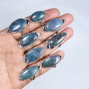 May include: A collection of gemstone rings with oval and teardrop-shaped stones. The stones are a mix of blue and green with red inclusions, set in silver-colored metal bands. The rings are displayed on a hand.