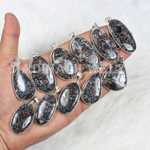 Peut inclure: Plusieurs pendentifs ovales et en forme de goutte d'eau avec une pierre mouchetée noire, blanche et rouge sertie dans du métal argenté. Les pendentifs sont présentés sur une surface blanche texturée.