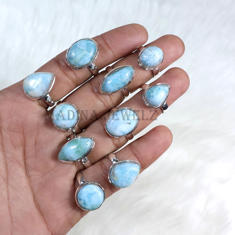 Pode incluir: Uma cole&ccedil;&atilde;o de an&eacute;is de prata com pedras de larimar azul claro em v&aacute;rias formas. Os an&eacute;is s&atilde;o exibidos em uma m&atilde;o, mostrando os diferentes cortes e engastes das pedras. As pedras preciosas t&ecirc;m uma apar&ecirc;ncia marmorizada.