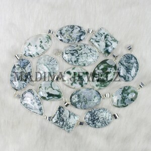 May include: A collection of gemstone pendants in various shapes, including oval, teardrop, and square. The pendants feature a white and green pattern, set in silver-colored metal. The image is taken on a white, textured surface.