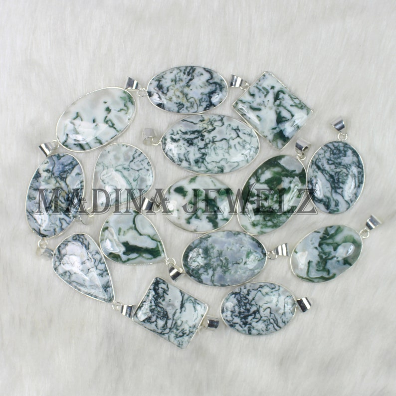 May include: A collection of gemstone pendants in various shapes, including oval, teardrop, and square. The pendants feature a white and green pattern, set in silver-colored metal. The image is taken on a white, textured surface.