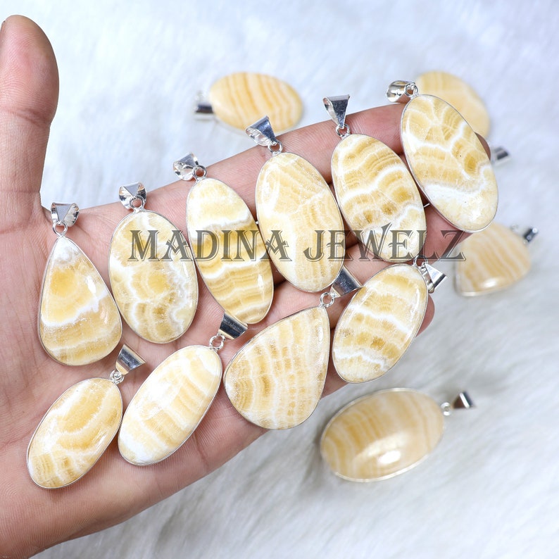 Pu&ograve; includere: Una collezione di pendenti lucidi, di forma ovale, con un'ansa argentata. I pendenti presentano un motivo a bande giallo crema e bianco. Il testo "MADINA JEWELZ" &egrave; visibile nell'immagine.
