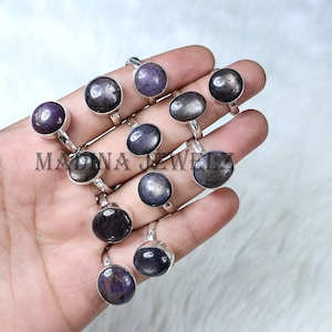 May include: A collection of gemstone rings with silver bands. The rings feature various dark-colored, oval-shaped stones, including shades of purple, gray, and black. The rings are displayed on a hand, showcasing the jewelry's design and color variations. The text "MADNA JEWELZ" is visible.