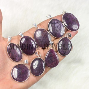 May include: A collection of oval and teardrop-shaped pendants with a deep purple gemstone set in silver. The pendants are displayed on a white surface. The text "MADINA JEWELLZ" is visible on some of the pendants.