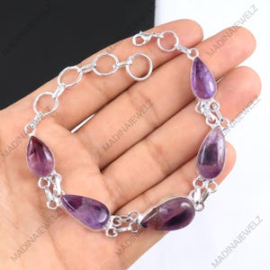 May include: A silver bracelet featuring teardrop-shaped amethyst stones. The purple stones are set in a silver chain with a lobster clasp closure. The bracelet is designed with a series of connected links.