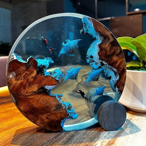 Könnte beinhalten: Ein rundes Kunstharz-Kunstwerk mit einem Holzsockel. Das Harz ist klar und enthält blaue und schwarze Mantarochen, einen Taucher und blaue und weiße Wellen. Der Holzsockel ist dunkelbraun mit einer natürlichen Holzmaserung.