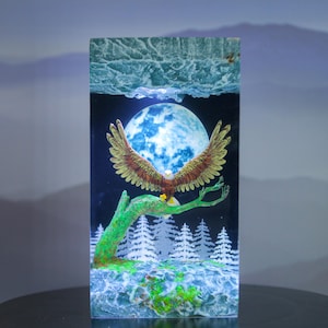 Puede incluir: Una pieza de arte de resina rectangular que representa un águila con las alas extendidas posada en una rama verde. El fondo incluye una luna llena, un bosque de árboles blancos y un cielo azul. El águila tiene plumas marrones y doradas.