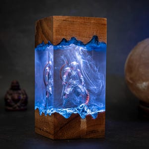 May include: A wooden lamp base with a clear resin insert containing a miniature figure of a warrior holding a bow and arrow. The resin is filled with blue and white swirls, creating a cloudy effect. The lamp base is lit from within, casting a blue glow.
