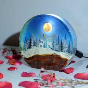 May include: A round resin night light featuring a winter scene. The scene depicts a snowy forest with two white deer standing beneath a full moon. The base of the night light is made of wood.