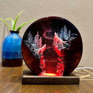 May include: A resin art piece with a circular wooden base. The resin is clear and contains a depiction of a forest scene with two white wolves howling at the moon. The resin is lit from behind with a red light, creating a glowing effect.