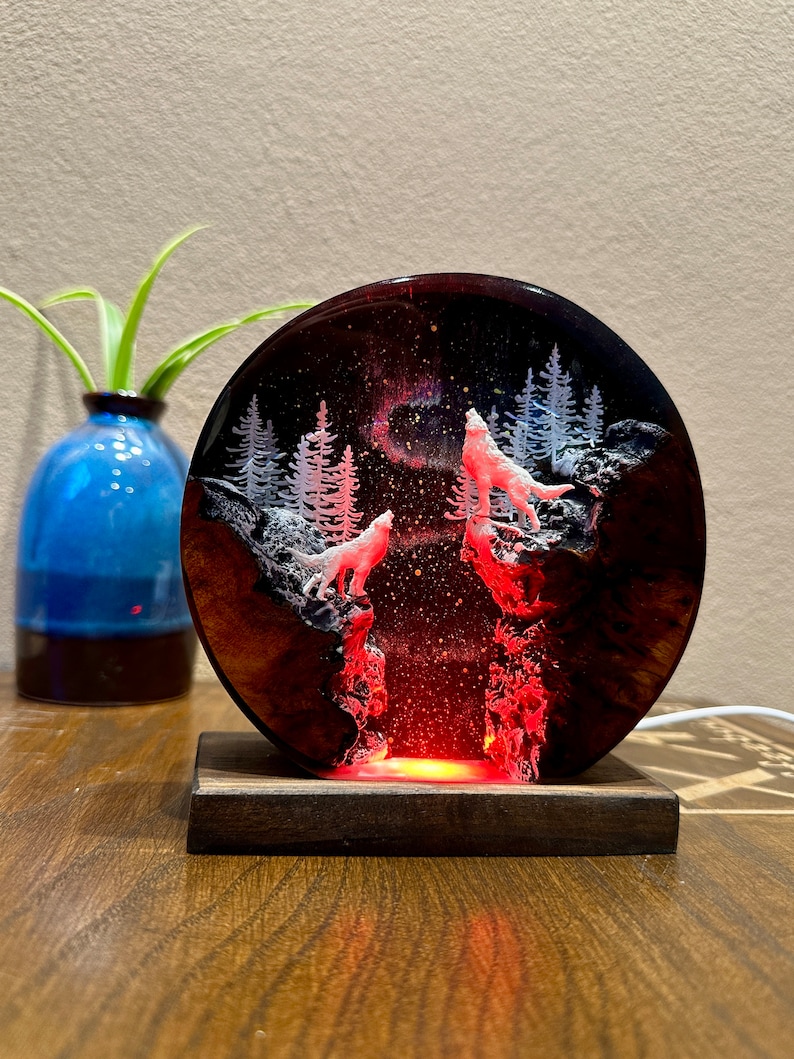 May include: A resin art piece with a circular wooden base. The resin is clear and contains a depiction of a forest scene with two white wolves howling at the moon. The resin is lit from behind with a red light, creating a glowing effect.