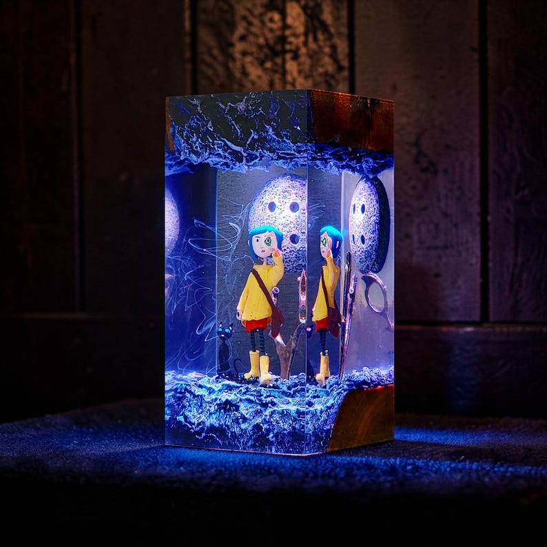 Puede incluir: Una pieza de arte de resina rectangular con una figura vestida de amarillo con cabello azul y un gato negro. La obra de arte est&aacute; iluminada con luz azul, mostrando una escena caprichosa con una esfera lunar y una base de madera.