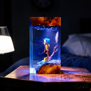 Puede incluir: Una pieza de arte de resina rectangular con una figura de pelo azul y abrigo amarillo. La obra de arte est&aacute; encerrada en resina transparente con base y parte superior de madera, y un fondo azul arremolinado.