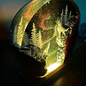 May include: A round, clear resin sculpture depicting a winter scene with two wolves howling at the moon. The sculpture is illuminated from below, casting a warm glow. The scene features a snowy forest, a starry sky, and a colourful aurora borealis.