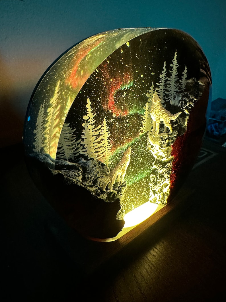 May include: A round, clear resin sculpture depicting a winter scene with two wolves howling at the moon. The sculpture is illuminated from below, casting a warm glow. The scene features a snowy forest, a starry sky, and a colourful aurora borealis.