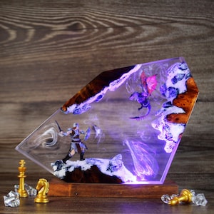 Puede incluir: Una escultura de resina de un guerrero luchando contra un dragón, colocada en un bloque de resina transparente con una base de madera. La escultura está iluminada desde abajo con luces LED moradas y blancas.