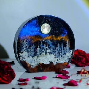 May include: A round resin art piece featuring a winter scene. The scene depicts a snowy forest with a full moon and two deer. The resin is clear and the scene is illuminated from behind.