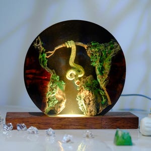 Könnte beinhalten: Eine runde Harzlampe mit einer grünen Schlange, die sich um einen Ast windet, vor einem Hintergrund aus grünem und braunem Moos. Die Lampe wird von innen beleuchtet und strahlt ein warmes Licht aus.