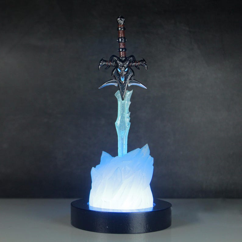 May include: A blue and silver sword with a skull-like handle is displayed on a black base with a blue light emanating from the base. The sword is on a white, frosted base that resembles ice.