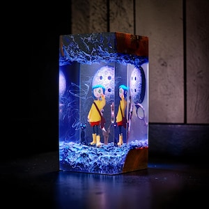Puede incluir: Una pieza de arte de resina rectangular que presenta una escena en miniatura con un personaje con abrigo amarillo y cabello azul. La escena incluye un gato negro, botones y una combinaci&oacute;n de colores azul y blanco. La parte superior e inferior son de madera oscura.