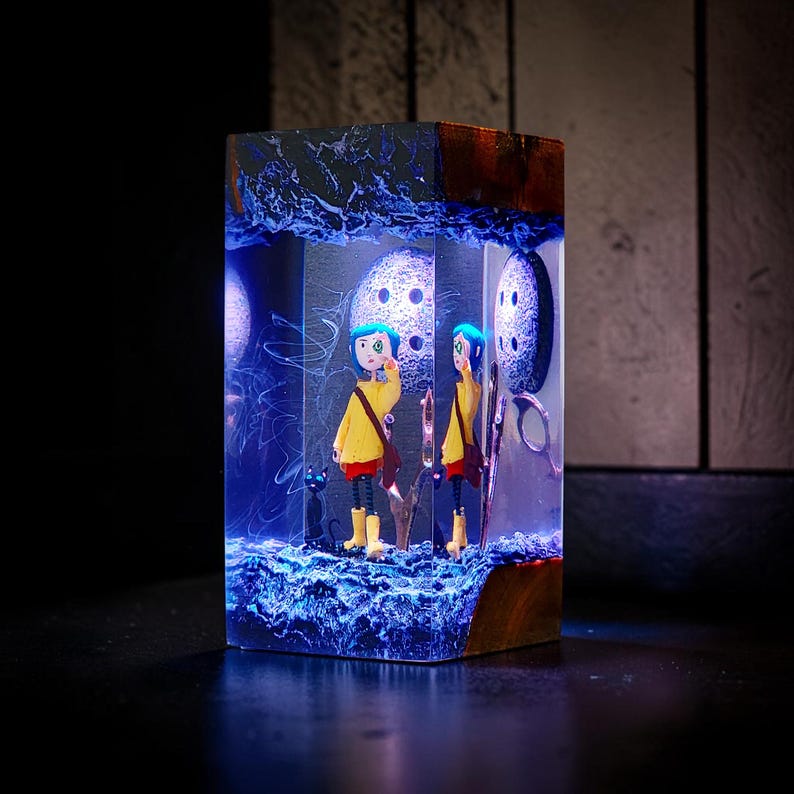 Puede incluir: Una pieza de arte de resina rectangular que presenta una escena en miniatura con un personaje con abrigo amarillo y cabello azul. La escena incluye un gato negro, botones y una combinaci&oacute;n de colores azul y blanco. La parte superior e inferior son de madera oscura.