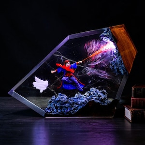 May include: A resin and wood diorama featuring a miniature figure in a blue robe and red scarf casting a glowing orange spell. A white owl figure flies nearby, set against a dark, swirling background with blue rocky textures encased in a clear geometric frame.