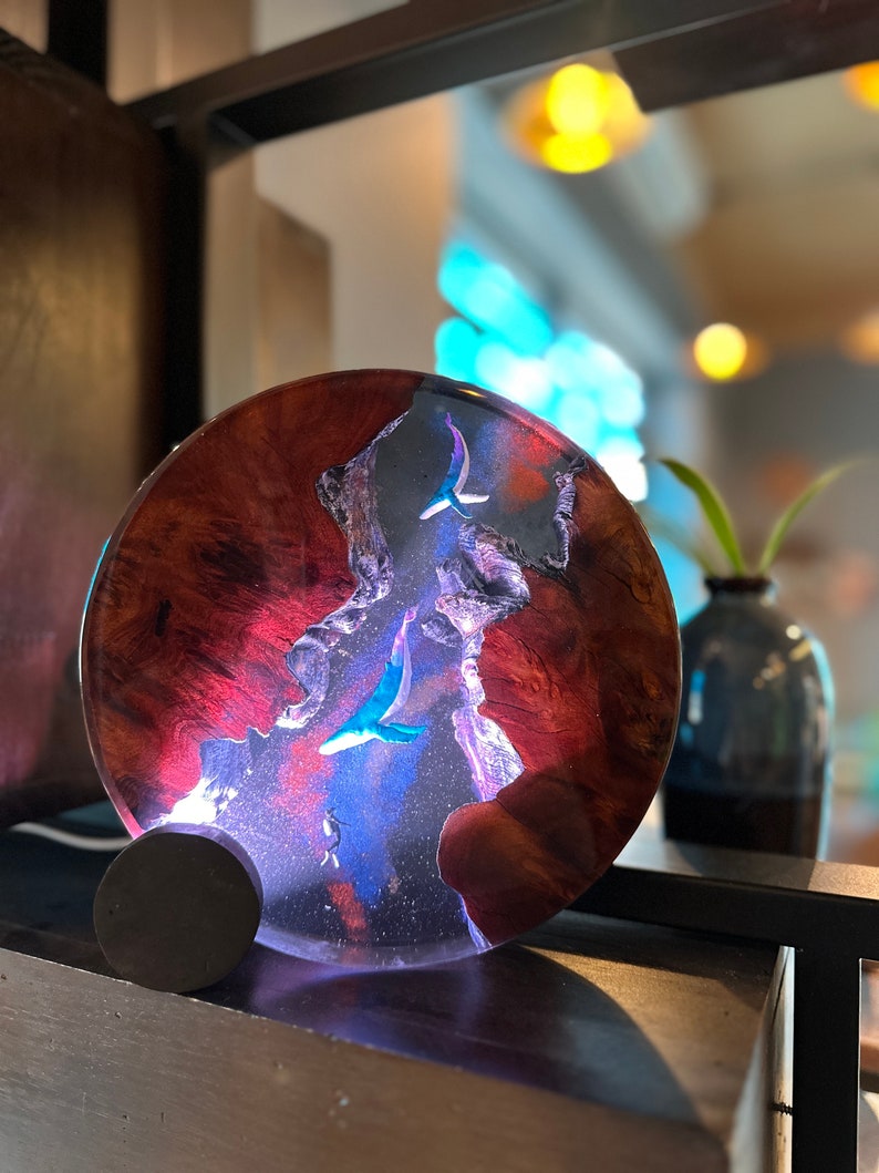 May include: A round, resin art piece with a dark wood base. The resin is clear and has a swirling pattern of blue, purple, and red. There are two blue whale silhouettes in the resin.