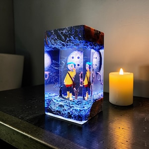 Puede incluir: Una pieza de arte de resina rectangular que presenta una escena con dos figuras con abrigos amarillos y un gato negro. La obra de arte est&aacute; iluminada con luz azul, con detalles de luna y botones. Una vela encendida est&aacute; cerca.