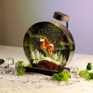 May include: A round resin sculpture featuring a glowing aurora borealis scene. Two orange foxes stand on a snowy hill with a forest behind them. The sculpture is illuminated by a small black lamp.