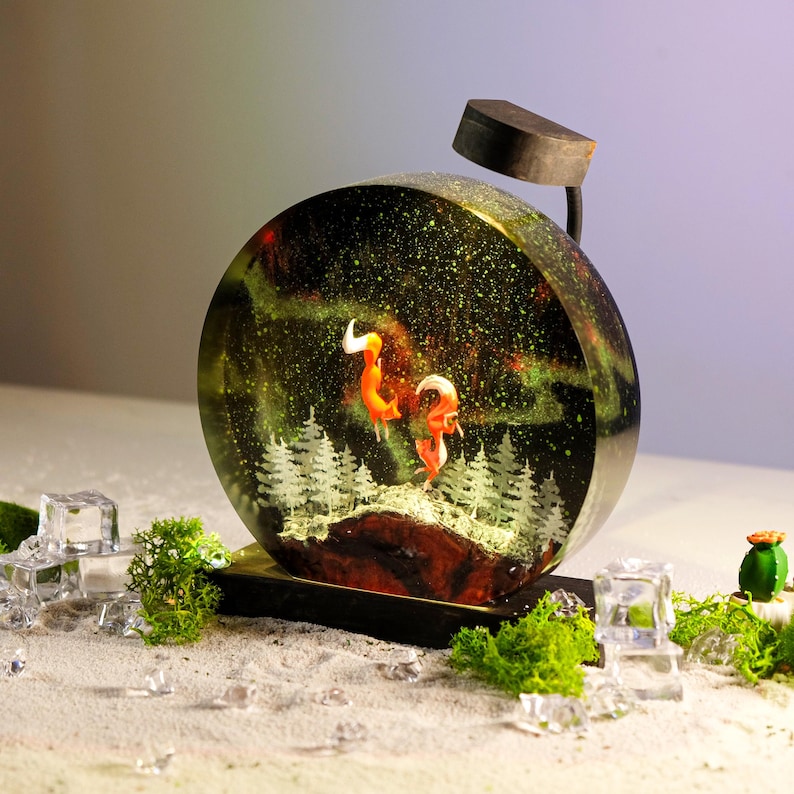 May include: A round resin sculpture featuring a glowing aurora borealis scene. Two orange foxes stand on a snowy hill with a forest behind them. The sculpture is illuminated by a small black lamp.