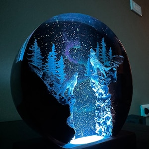 May include: A glass dome with a glowing blue light inside. The dome contains a scene of two wolves howling at the moon, surrounded by trees and a starry sky.