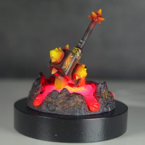 May include: A black and gold hammer with spikes, resting on a lava-like base with red glowing lights. The hammer is on a black circular base.