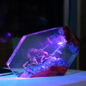 May include: A clear resin sculpture of a skeletal alien creature with a purple glow emanating from its chest. The sculpture is set on a wooden base and encased in a clear resin block.