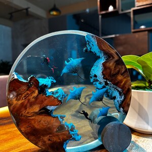 May include: A round resin art piece with a wooden base. The resin is clear and contains blue and white manta rays, a diver, and a blue and white wave pattern. The wooden base is dark brown with a natural wood grain.