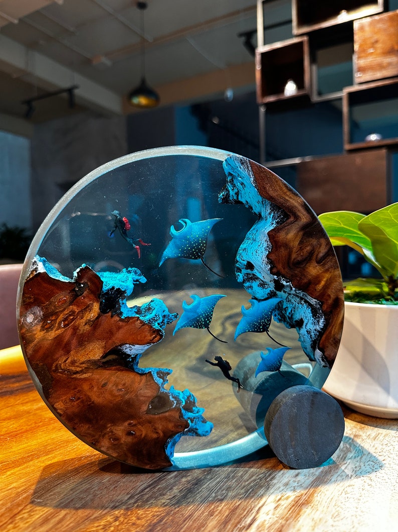 May include: A round resin art piece with a wooden base. The resin is clear and contains blue and white manta rays, a diver, and a blue and white wave pattern. The wooden base is dark brown with a natural wood grain.
